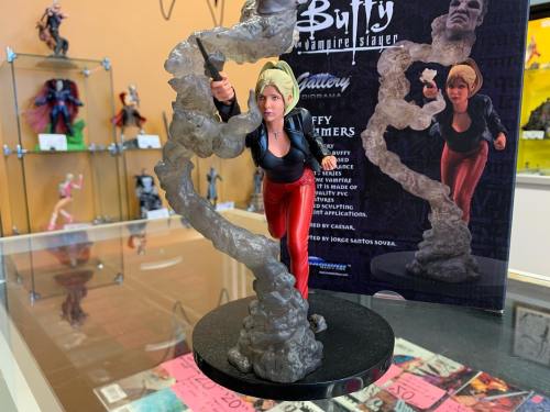 A figurine of Buffy Summers in action, surrounded by a swirling gray effect, displayed with promotional materials.