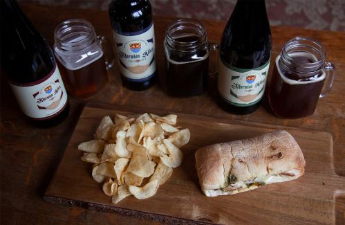 A wooden board with a sandwich, potato chips, and several bottles of beer in the background.
