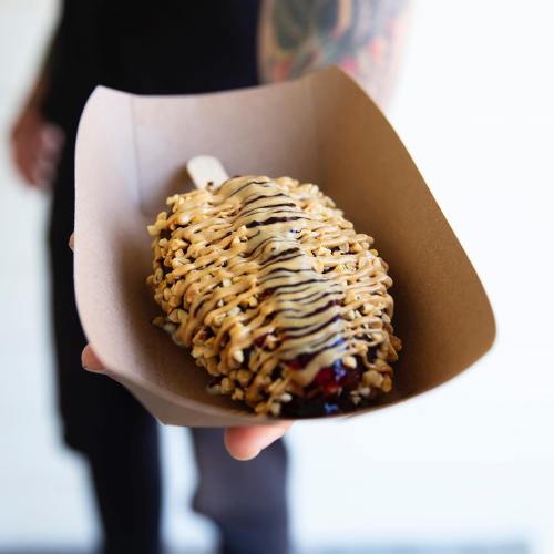 A hand holds a dessert on a stick, drizzled with chocolate and topped with crushed peanuts, in a brown paper tray.