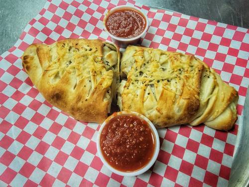Two calzones on a red and white checkered paper, served with two small cups of marinara sauce.