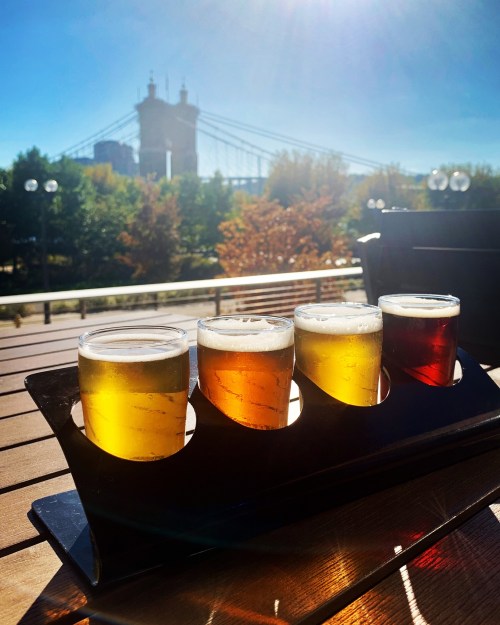 Four beer samples in a flight on a wooden table, with a sunny park and bridge in the background.