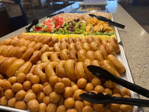 A buffet table featuring a variety of donuts, fruit, and pastries, with tongs for serving.