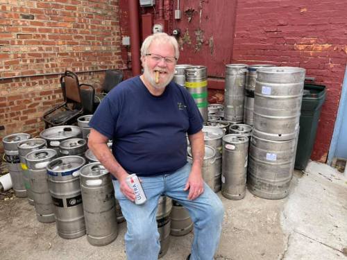 A man with glasses and a beard sits among metal kegs, holding a can, in a brick alleyway.