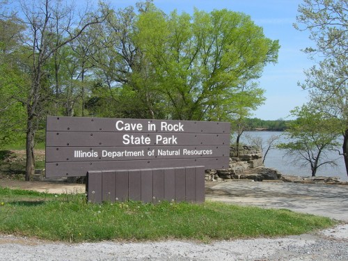 Sign for Cave in Rock State Park, with trees and a river in the background on a clear day.