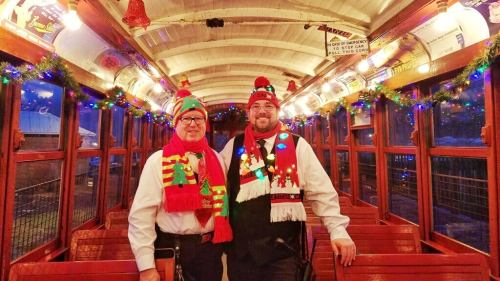 Two men in festive attire stand inside a decorated train car, adorned with Christmas lights and holiday decorations.