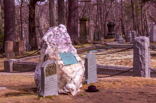 A rocky memorial in a cemetery, surrounded by gravestones and trees, with a hat resting on the ground nearby.