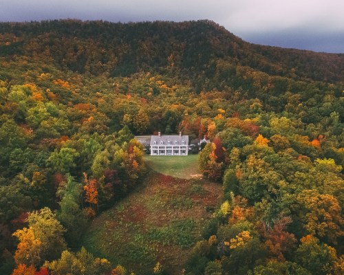 Aerial view of a large house surrounded by vibrant autumn foliage and rolling hills under a cloudy sky.