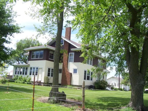 A large two-story house with a mix of brown and cream siding, surrounded by trees and a green lawn.