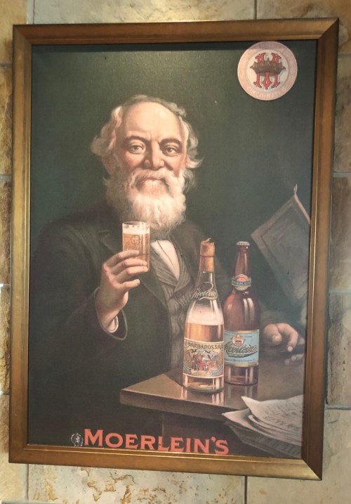 Vintage advertisement featuring a bearded man holding a beer, surrounded by bottles and a newspaper, framed on a wall.