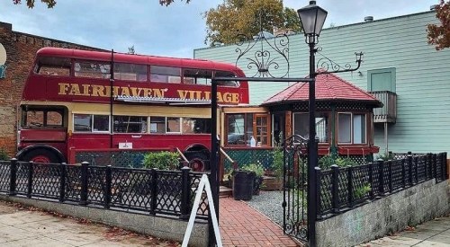 A red double-decker bus converted into a building, surrounded by a garden and a decorative fence in a village setting.