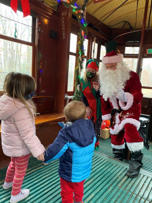 Children interacting with Santa and an elf on a decorated trolley, all wearing festive attire and masks.