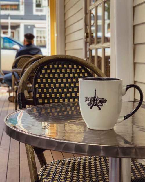 A coffee mug with "Mornings in Paris" sits on a table, with a person in the background enjoying the outdoor setting.