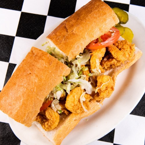 A sandwich filled with fried shrimp, lettuce, and tomato, served on a toasted bun with pickles on the side.