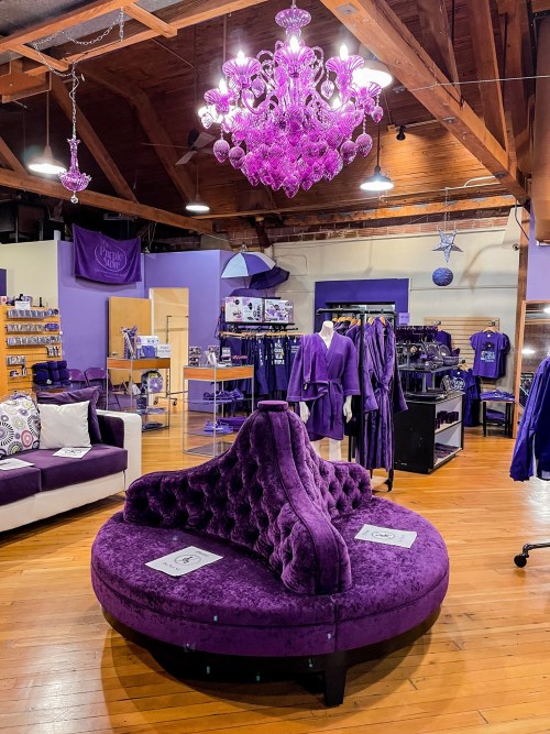 A cozy store interior featuring a large purple velvet sofa, a chandelier, and various purple-themed merchandise.
