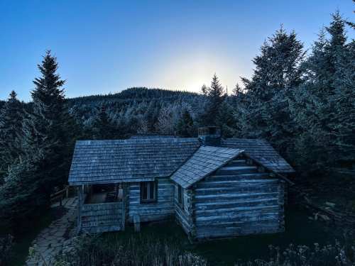 A rustic log cabin surrounded by tall evergreen trees, set against a backdrop of a mountain at dusk.