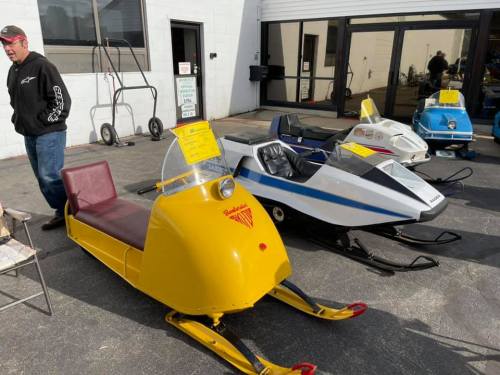 A man stands beside vintage snowmobiles in various colors, displayed outside a building.