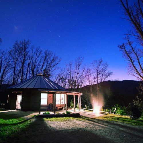 A cozy cabin surrounded by trees under a starry sky at dusk, with a warm glow from a fire pit in the foreground.