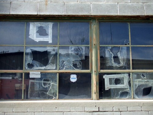 A weathered building window with multiple broken panes and scattered paper remnants inside.