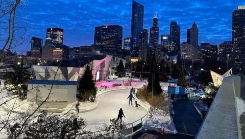 A winter scene with people ice skating in a park, surrounded by a city skyline at dusk.