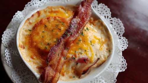 A creamy dish topped with melted cheese, garnished with herbs, and accompanied by a crispy strip of bacon.