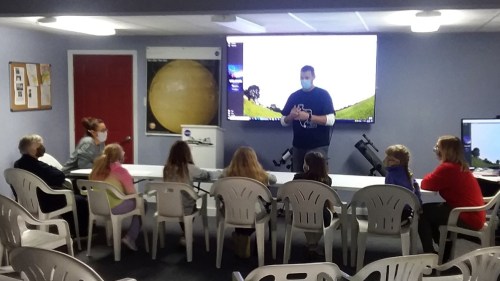 A group of children and adults sit at a table in a classroom, listening to a speaker in front of a large screen.