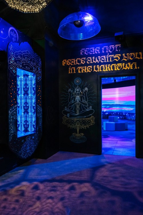 A dimly lit room with blue lighting, featuring a decorative wall and a doorway leading to a scenic view.