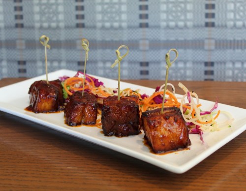 Four glazed cubes of grilled meat on skewers, served with colorful shredded vegetables on a white plate.