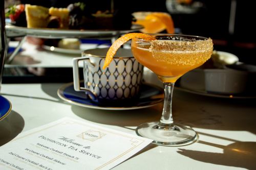 A cocktail with an orange twist sits beside a decorative cup and a menu on a table, illuminated by natural light.