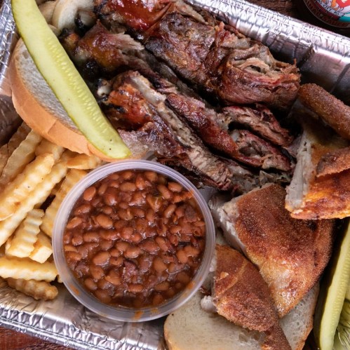 A tray of barbecue ribs, pickles, fries, baked beans, and slices of bread.