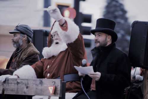 Santa Claus waves from a float, accompanied by two men in winter attire, during a festive parade.