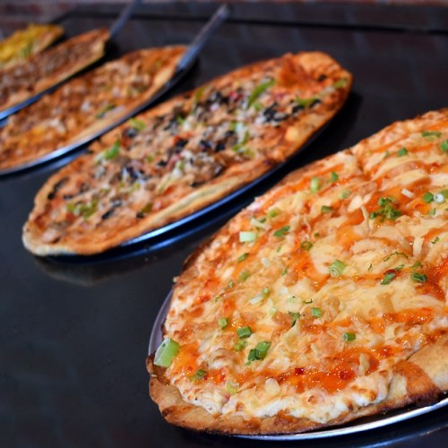 A variety of pizzas on metal trays, featuring different toppings and crust styles, arranged in a row.