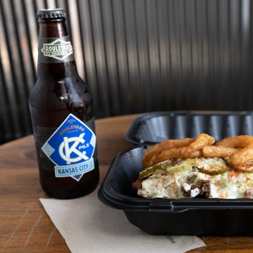 A bottle of Boulevard beer next to a takeout container filled with food on a wooden table.
