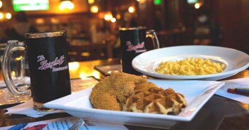 A plate with fried chicken and waffles, alongside a bowl of macaroni and cheese, with two mugs on the table.