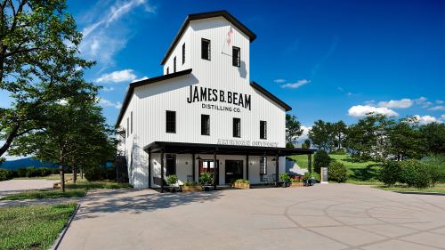 A white distillery building with "James B. Beam Distilling Co." sign, surrounded by greenery and a clear blue sky.