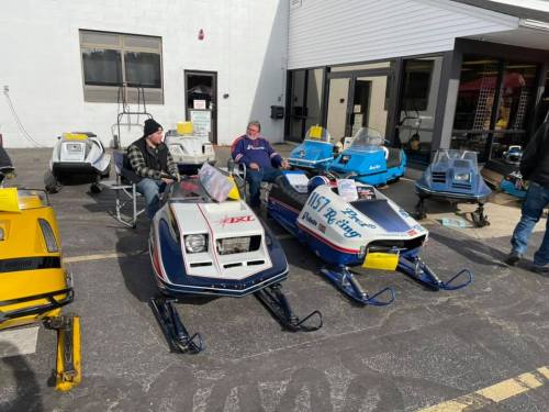 Two men sit among vintage snowmobiles displayed outside a building, with various models lined up in the background.