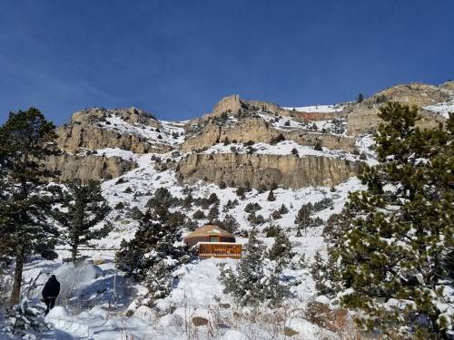 A snowy landscape with a cabin nestled among trees, set against towering rocky cliffs under a clear blue sky.