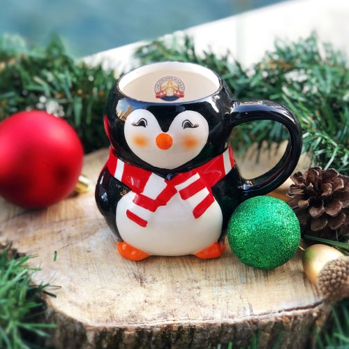 A cute penguin-shaped mug with a red scarf, surrounded by festive decorations like ornaments and pinecones.