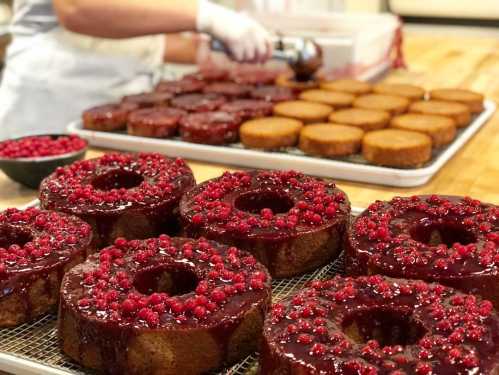 A variety of cakes topped with red berries and glaze, arranged on trays in a bakery setting.