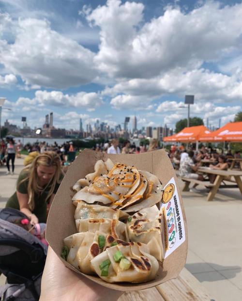 A hand holding a container of dumplings with a city skyline and people in the background on a sunny day.