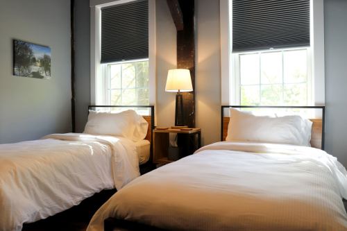 A cozy bedroom with two twin beds, a bedside table, and a lamp, featuring large windows with blinds.