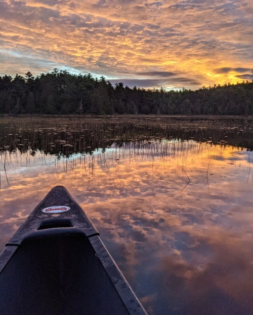 A canoe's bow on a calm lake reflecting a colorful sunset and surrounding trees.