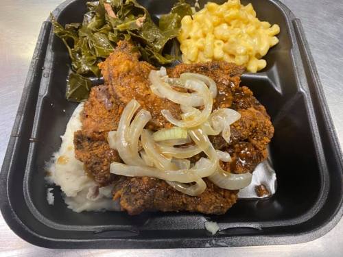 A black takeout container with fried chicken topped with onions, collard greens, macaroni and cheese, and mashed potatoes.