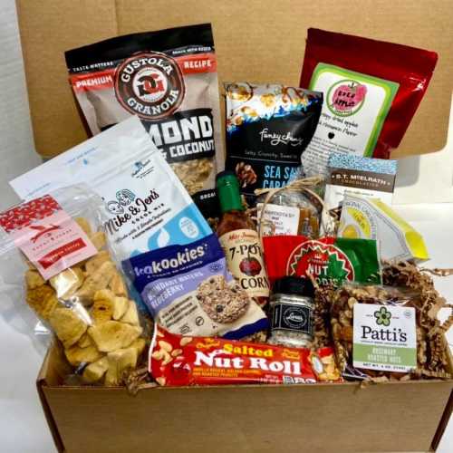 A gift box filled with various snacks, including granola, cookies, popcorn, and sauces, arranged with decorative filler.