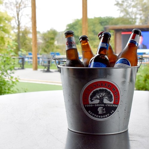 A bucket filled with various beer bottles on a table, with a scenic outdoor setting in the background.