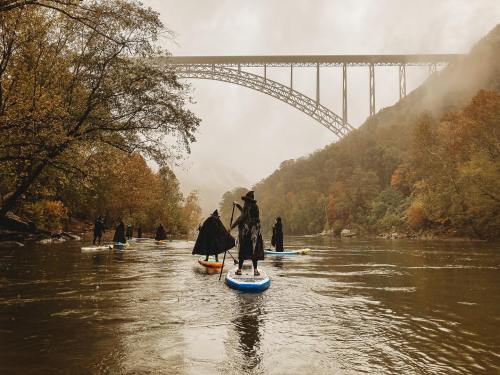 A group of people in dark cloaks paddle on a river under a large bridge, surrounded by autumn foliage and mist.