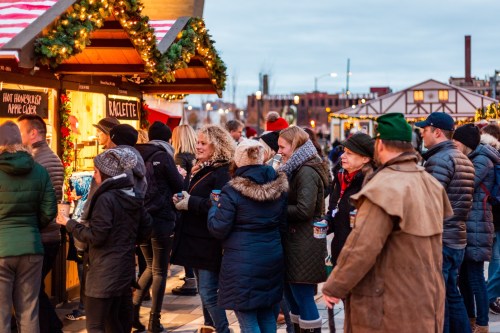 A festive crowd gathers at a holiday market, enjoying food and drinks under twinkling lights and decorations.