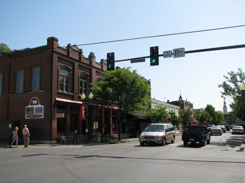 A sunny street scene with shops, trees, and traffic lights, featuring pedestrians and parked cars.