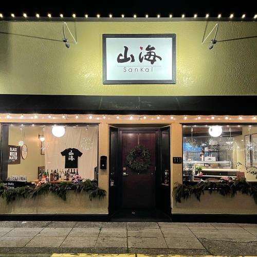 A cozy restaurant exterior with warm lighting, featuring the name "Sankai" and festive decorations.