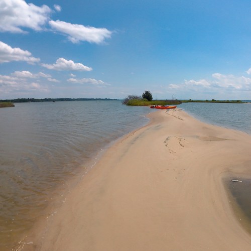 A sandy path leads to calm waters under a blue sky, with a kayak resting nearby and greenery in the distance.