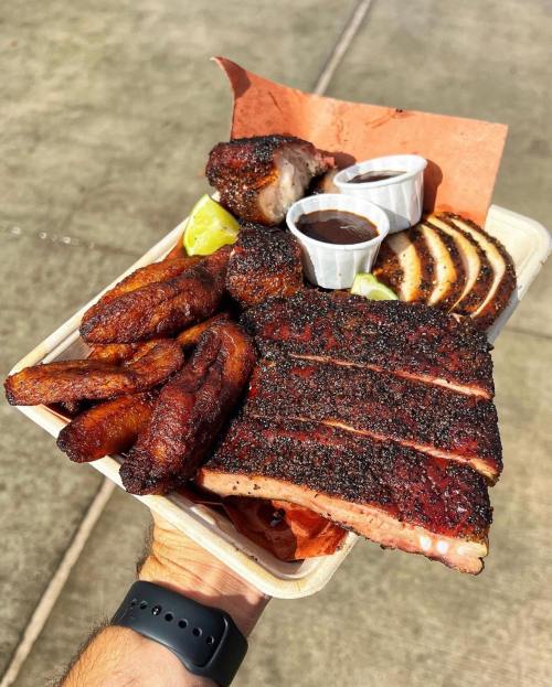 A hand holding a tray of smoked meats, including ribs, sausage, and plantains, with dipping sauces and lime wedges.